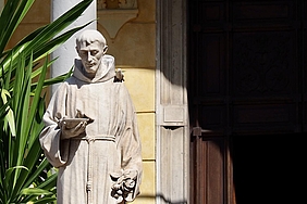 Steinstatue eines Mönchs mit Buch in sonnigem Hof neben einer Pflanze