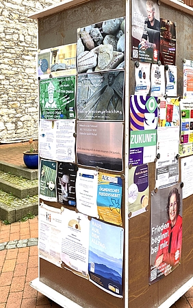 Säule mit vielen bunten Plakaten und Flyern im Außenbereich vor einer Steinmauer. 