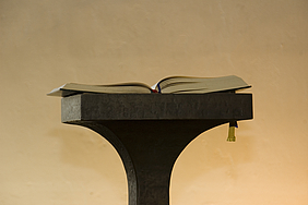 Aufgeschlagenes Buch auf einem Pult vor beigefarbenem Hintergrund.