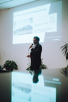 Mann mit Mikrofon; im Hintergrund ein großes Bild an der Wand