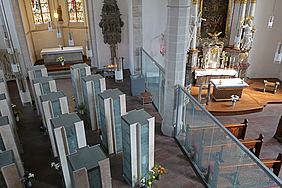 Heller Kirchenraum mit Altären und modernen Stelen, bunte Glasfenster im Hintergrund.
