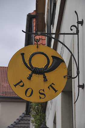 Gelbes Post-Schild mit schwarzem Posthorn an einer Hauswand.