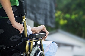 Ältere Person sitzt im Rollstuhl, Assistenzperson steht daneben.