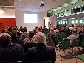 Sitzende Personen in einer Halle bei einem Vortrag